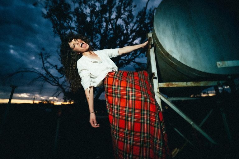Australian country singer Fanny Lumsden leaning playfully against a water tank at twilight in a white blouse and red tartan skirt with tongue out.